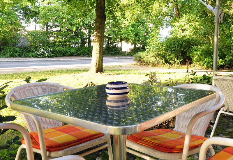 Ein silberner Tisch mit vier Stühlen im Vordergrund, Blick auf Baumstamm mittig und Busch rechtsseitig, im Hintergrund verläuft eine Straße